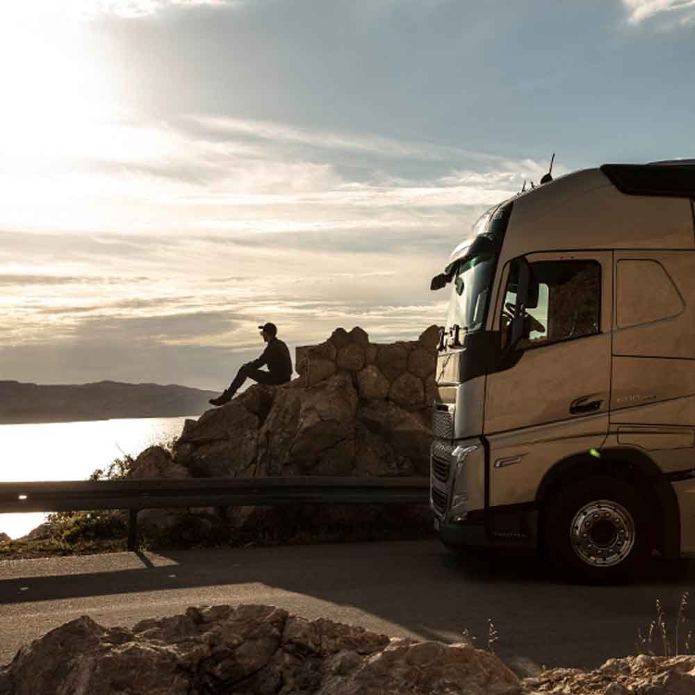 Un conducteur est assis sur un rocher face au coucher de soleil sur la mer, avec son Volvo FH garé à côté