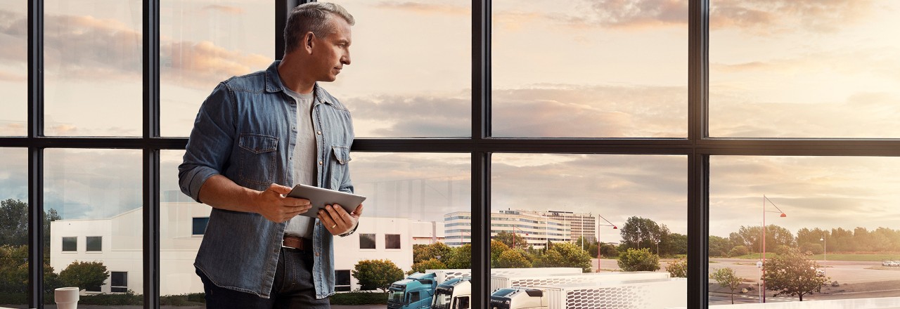 Un homme tenant une tablette se tient devant une fenêtre et regarde son parc de véhicules